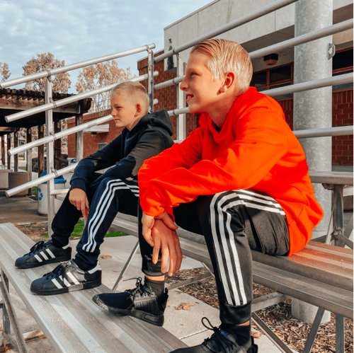 2 young boys wearing red and black hoodies and adidas sweatpants with black tennis shoes on bleachers